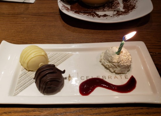 A rectangular plate with one round white chocolate, one round dark chocolate, a curved stripe of berry sauce, and a lit candle standing in a mound of whipped cream the same size as each candy.