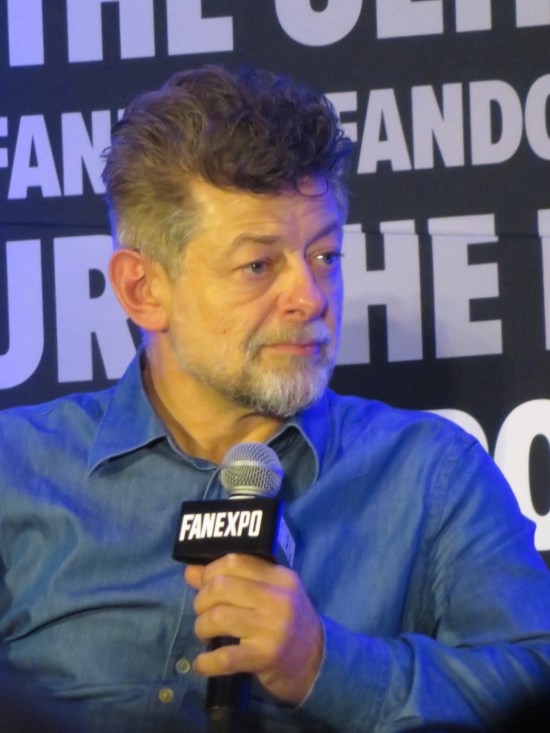 Andy Serkis sitting on stage, holding a mic, listening to his host intently.
