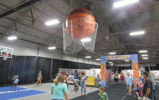 A pavilion with exhibits and kids running around. A giant inflatable basketball looms overhead inside a matching basket.