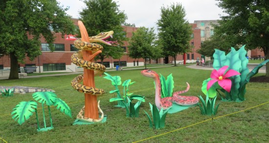 Two big snake sculptures on a lawn. One is on a green tarp, the other coiled around a fake tree with no branches. Fake aquamarine plants surround them.