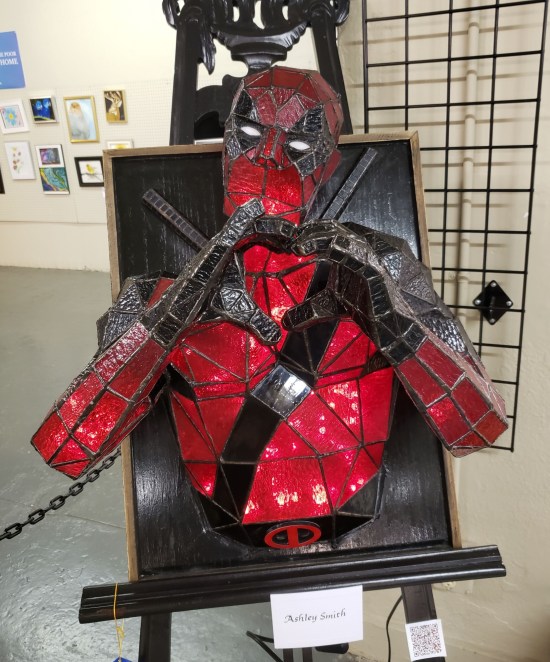 A quasi-stained glass sculpture of Deadpool's upper half. He has his swords on his back. His hands are cupped in a heart shape.