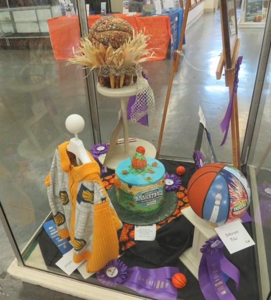 A tall vitrine containing a homemade Pacers sweater, two Indiana-themed basketballs, and a basketball top hat.