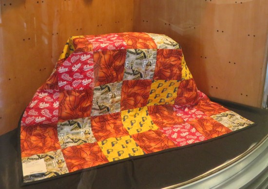 A red-and-orange Pacers quilt draped over a box in a display case.