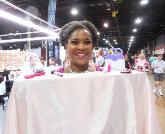 A woman wearing a table with her head poking through a hole in the middle. Hairstyling equipment festoons the tabletop around her.