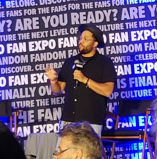 Victor Dandridge Jr on a Fan Expo stage with a mic and hat.
