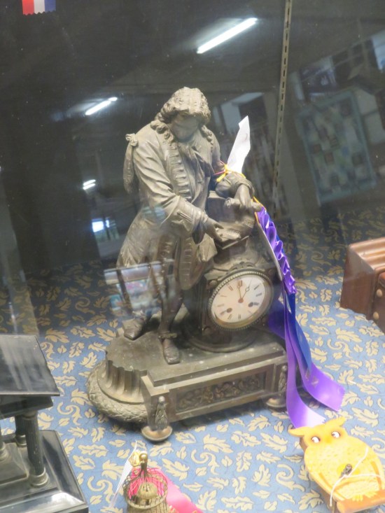 A tiny scale model of one of those clocks they used to mount on the ledges outside big-city department stores. Next to it is a scale model of a Founding Father I don't recognize. They share a base. They're entirely metal. It's in a vitrine with other antiques. It was awarded a purple Grand Prize ribbon.