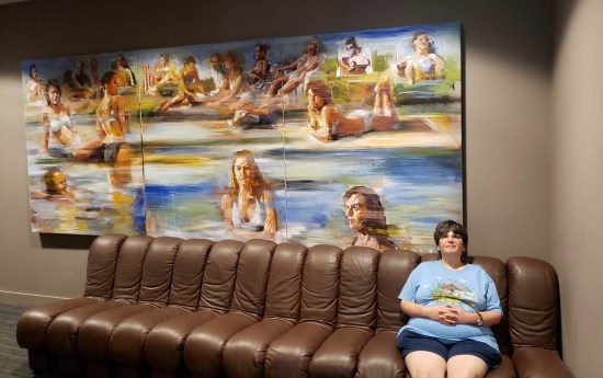 Anne sitting on one end of a very long brown couch. On the wall above is a mural of numerous painted bikini babes.