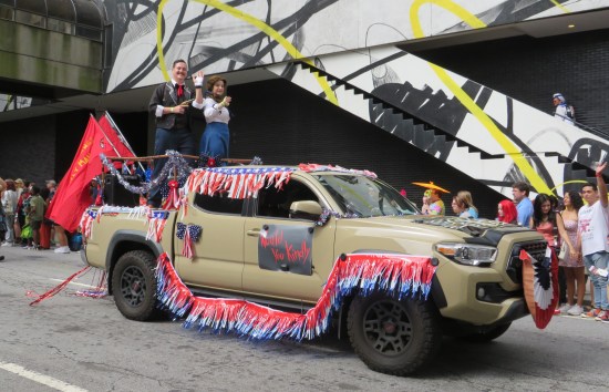 A 1920s couple stands up in the back of a beige pickup truck festooned with Fourth of July decorations. The duo waves and smiles to us. Red flags hang from the back with the Bioshock catchphrase "No gods or kings, only man."