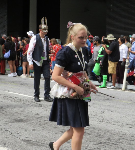 Creepy little girl with a big hydraulic needle and heavy eye shadow. In the background, tattered house servant in a bloodied bunny mask.