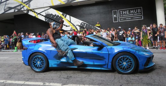 A blue sports convertible with thick black outlines painted all over, apropos of the Borderlands video games' visual aesthetic. A guy in a hockey mask sits on the back holding a weaponized buzzsaw, one leg hanging off the side.