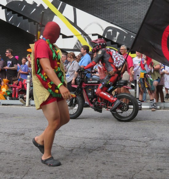 Deadpool cosplayer in tiny shorts with a large Mexican tortilla and fake ingredients wrapped around him. Another Deadpool cosplayer rides past on a motorcycle.