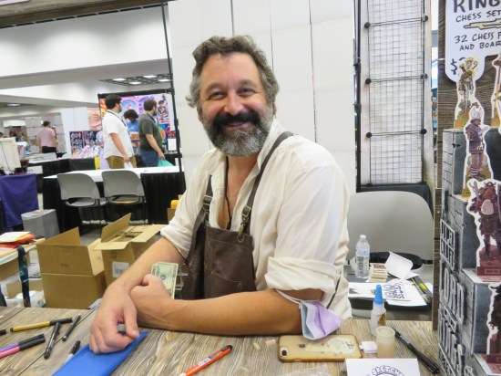 Chris Schweizer sitting at his table in a brown workshop apron, smiling and surrounded with examples of what he does.