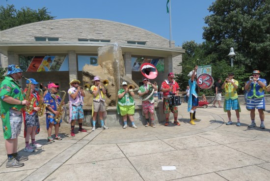 Eleven musicians in a curved row, playing in front of a beige building with a 10-foot rock fountain in front of it. Three saxes, two trumpets, three trombones, one tuba, one snare drum and one bass drum.