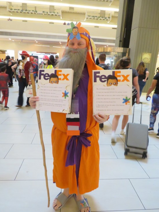 cosplay: guy in orange robes carrying two Federal Express envelopes, each bearing pics of the top half of a guy's head with googly-eyes. Robed guy also has the full head photo with googly-eyes on his own forehead.