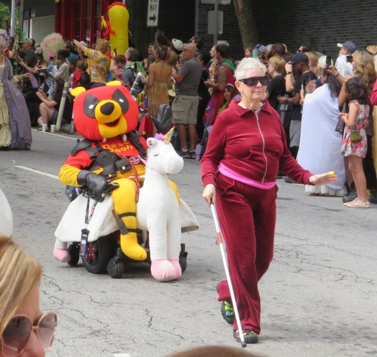 Winnie the Pooh cosplayer also wearing Deadpool costume, in motorized wheelchair with unicorn trimmings. In front of him is a blind elderly woman in a red tracksuit.