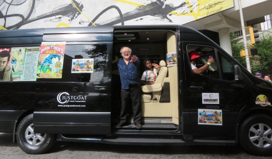 Comics writer Don McGregor in the back of a black van, standing and waving from its open side door. Black Panther panels decorate the outside.