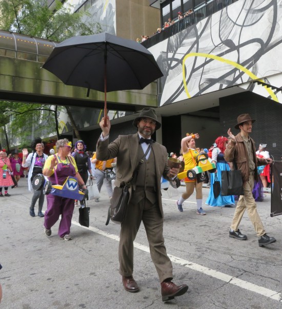 cosplay Sean Connery from "Indiana Jones and the Last Crusade" walks and looks at the camera while holding his big open umbrella aloft.