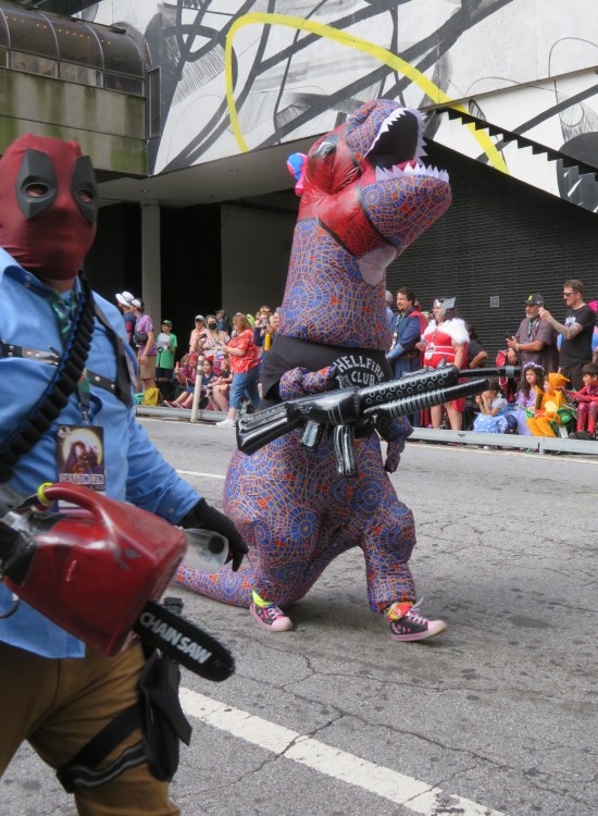 Foreground: a Deadpool cosplayer also wearing a costume of Ash from "Evil Dead", including chainsaw hand. Behind him, an inflatable T-Rex with Marriott Carpet skin wears a Deadpool mask and a black Stranger Things Hellfire Club T-shirt, and carries an inflatable AK-47.