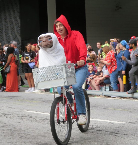 cosplay: adult Elliott on a bicycle with a blanketed E.T. puppet in the front basket