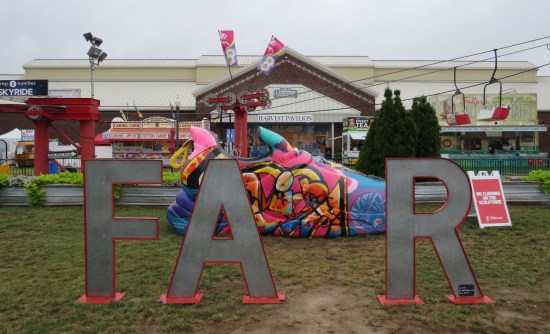 Big letters F, A, and R stand in grass, with a gap where you're supposed to stand like an I and spell "FAIR". Behind the letters is a giant tennis shoe with stylized faux graffiti. Farther back is the Harvest Pavilion building.