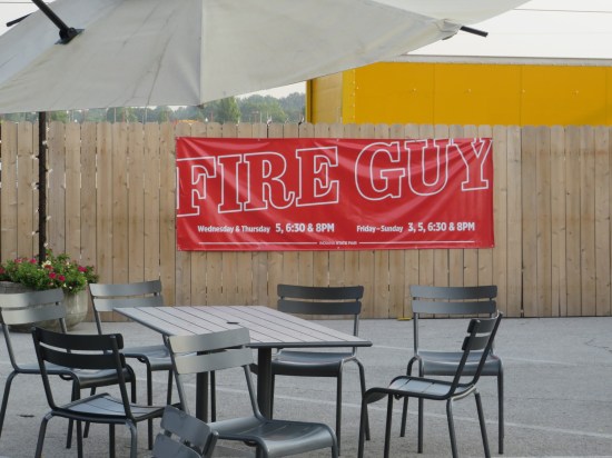Red banner hung on a wooden fence advertising a performer named FIRE GUY, who did fire stunts five days a week during the fair.
