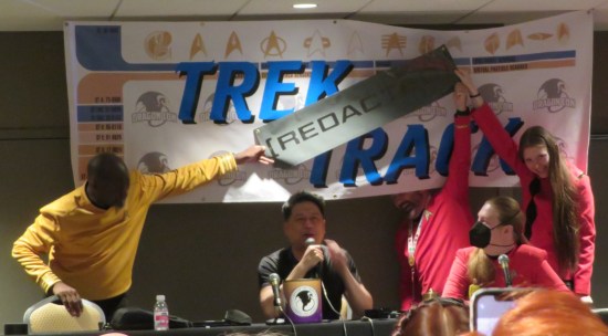 Garrett Wang trying to speak from his table to our audience. Behind him hangs a big "Trek Track" banner. For a few fun minutes his crew covered the word "Trek" with a smaller banner that says "REDACTED".