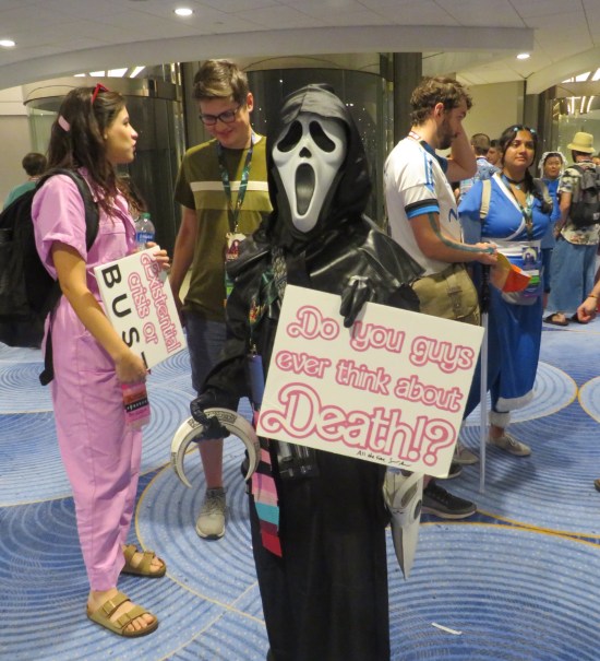 cosplay: Ghostface from "Scream" with a sign in Barbie font: "Do you guys ever think about death?"