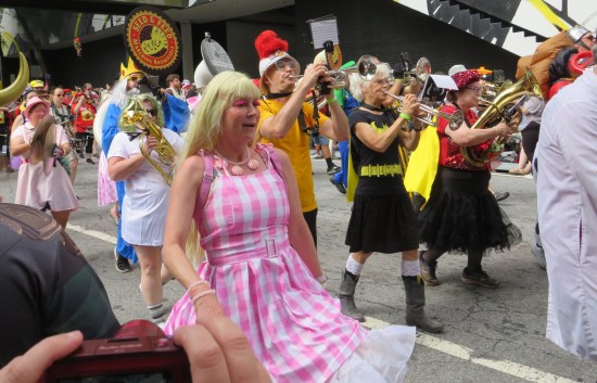 Gingham Barbie alongside a wacky marching band that includes a Batgirl.