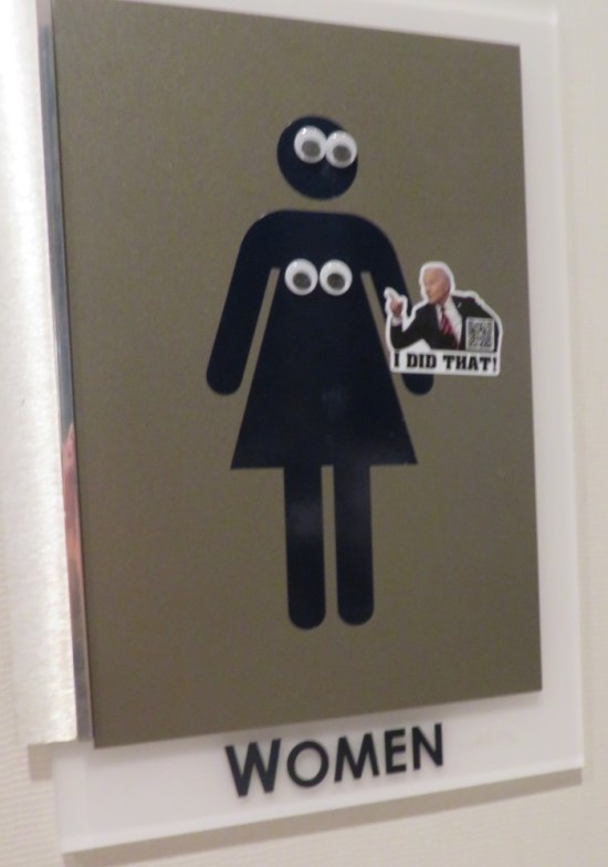 women's restroom sign with googly-eye stickers on the female figure's face and flat chest. A Joe Biden sticker is randomly stuck to her left arm.