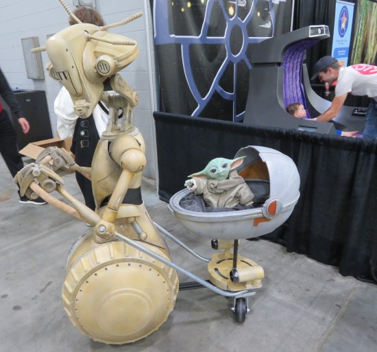 Grogu and Rickshaw Droid models. Behind them, a real father places his real baby in a full-size replica of Palpatine's throne.
