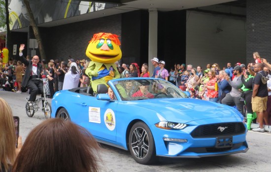 Someone in a big stuffed H.R. Pufnstuf costume riding on the back of a convertible with a very tiny and old Marty Krofft barely visible in the front passenger seat.