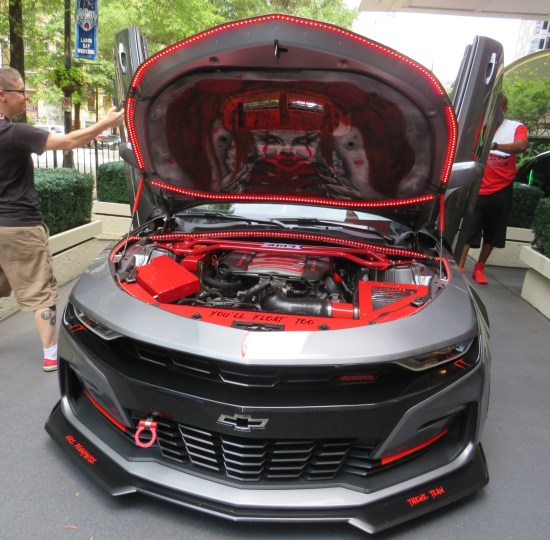 The Pennywise car parked, with hood open. A Pennywise head is painted inside the hood. Down on the engine block is written the words "You'll float too."