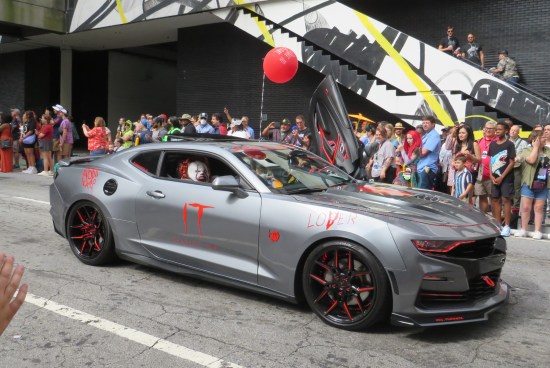 A car tricked out with logos from Stephen King's "It". Two Pennywise cosplayers ride inside. A red helium balloon is attached to the roof.