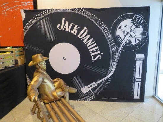 A statue of Jack Daniels sitting on a bench, turned to face a corner that had a large Jack Daniel's logo on a picture of a vinyl turntable.