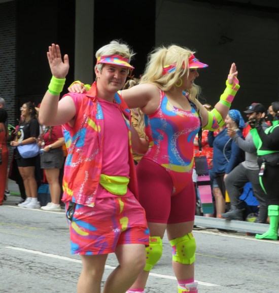 Movie Ken and Barbie walking together, all in pink. Barbie faces away from us, Ken toward us, waving.