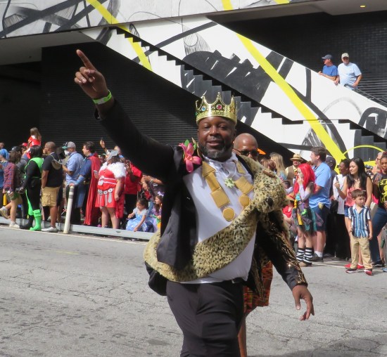 cosplay: imitation James Earl Jones in gold brown, totem necklaces, and fake leopard-skin sash. The king points diagonally to the sky.
