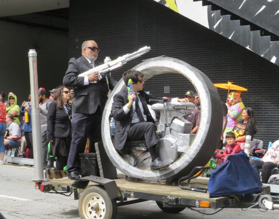 Two Men in Black hang onto a wheel-shaped cycle, set on a flatbed hitched to an offscreen car.