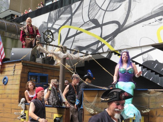At least eight pirates and one mermaid aboard a rolling pirate ship. The mermaid and one pirate are smiling directly into the camera.