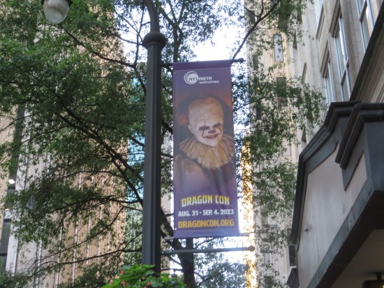A Dragon Con banner with a Pennywise cosplayer on it, hung un a high light pole.