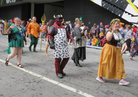 Deadpool wearing a baseball cap with cattle horns and an apron covered in cat silhouettes. By his side are cosplayers doing Link from "Legend of Zelda", Aquaman, and others.
