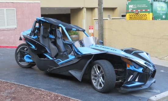 A blue Polaris Slingshot parked on asphalt -- not on the street, but on the other side of the sidewalk.
