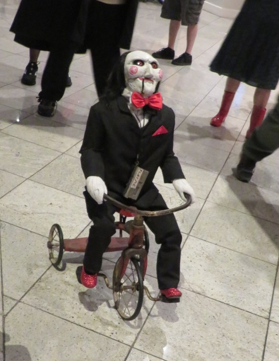 A Jigsaw puppet from the "Saw" films, rolling around on a remote-control tricycle.
