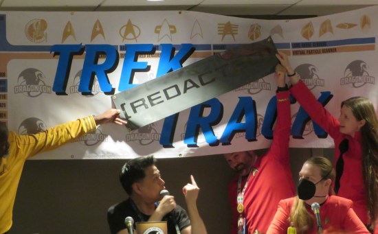 Garrett Wang sitting at a table at the front of a panel room, pointing at the Trek Track banner hanging behind him. Folks in classic Trek uniforms are jokingly covering "TREK" with a big "REDACTED" sticker.