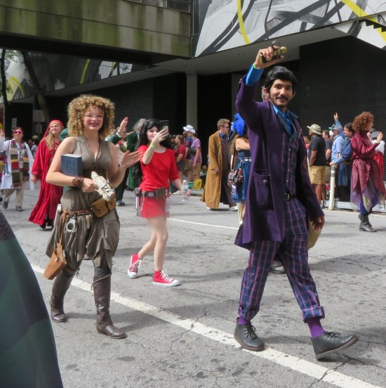 River Song with white sci-fi gun, waving with her free hand and a thick book tucked under the other arm. The Master is wearing all purple and bearing a fancy detonator.