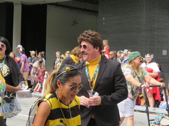 Robert Goulet cosplayer swings around with a martini. A woman in a bee costume looks away and marches onward.