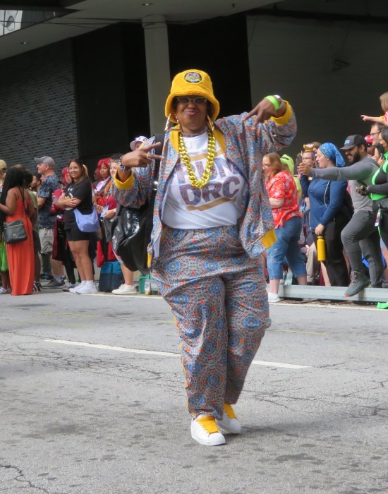 a lady dressed like the guys in Run-DMC with yellow Kangol hat, fake gold necklaces, and Cult of Marriott Carpet jacket and pants.