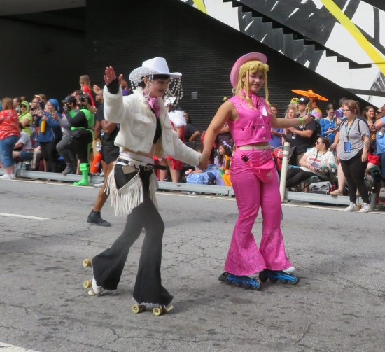 Movie Barbie in pink cowboy gear, holding hands with another young lady in non-Barbie non-pink cowboy gear -- white hat, jacket, and frills hanging from her belt. Both are on roller skates.