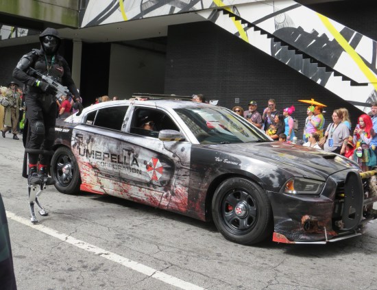 Heavily damaged police car with the Umbrella Corporation fictional logo on the door. A black-clad, play-armed security cosplayer on kangaroo shoes walks next to it.