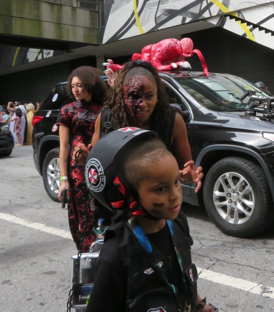 kid in an Umbrella Corporation soldier costume walking nonchalantly as if unaware that behind him, a Black woman in zombie makeup prepares to devour him.