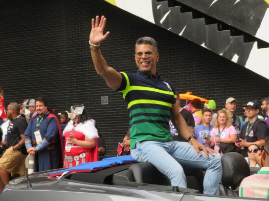 Wilson Cruz sitting on the back of a moving convertible. He smiles, waves, and wears a blue, yellow, and green-striped top, and two bracelets on his waving arm.